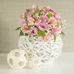 On a clean white surface that could easily be a Grove Park kitchen island or hallway console, a generous floral centerpiece radiates soft colour and calm, framed by a subtly textured light beige wall that keeps all focus on the flowers. At the heart of the scene is a striking white vase, rounded and slightly bulbous, formed from white strips interlaced into an open, lattice-style pattern, giving it a handcrafted, contemporary-rustic character. It feels solid yet airy, like a sculptural basket reimagined for modern interiors around Chiswick or Lee. Overflowing from this vessel is a carefully balanced bouquet of pastel pink and lavender roses, their petals velvety and lush, with some blooms fully open and others just beginning to unfurl, suggesting movement and life. Between them, elegant pink alstroemeria lift their slim, trumpet-shaped flowers, each petal brushed with fine streaks of deeper pink or soft golden tones at the throat, lending the bouquet a painterly detail best appreciated up close. Punctuating the soft pastels are bright lime green button chrysanthemums, compact and spherical, adding playful contrast and energy while drawing the eye across the arrangement. Delicate clusters of white baby's breath weave through everything, like tiny clouds suspended between stems, creating a gentle halo around the main blooms. Deep green foliage is tucked throughout, adding structure, freshness, and a hint of woodland fragrance. To the left of the vase rest two decorative spheres fashioned from natural-toned, woven strips, echoing the pattern of the vase and adding balance and rhythm to the composition. The overall impression is of quiet celebration and refined taste: a piece that could brighten a Grove Park living room for an anniversary, a new home, or a softly spoken thank you, arranged with the considered touch of a local florist who understands how flowers can change the mood of a room.