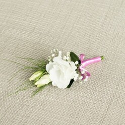 A refined floral boutonnière is photographed from above, resting at a gentle diagonal on a warm, light beige fabric that looks like delicately woven linen, its subtle texture echoing the relaxed elegance of homes around Grove Park. At the centre of the design is a single, fully opened white lisianthus flower, its ruffled petals layered generously, creating a soft, rounded rosette with a faint creamy glow at the throat. Just to the left sit two unopened lisianthus buds, slender and tapered, their pale green and ivory tones suggesting new beginnings, the sort of symbolism couples often ask for when we design wedding pieces at Grove Park Florist. Surrounding the trio of blooms are tiny clouds of gypsophila, or baby's breath, each small white floret hovering on fine stems to give a light, airy fullness without overwhelming the main flower. From behind the lisianthus, slim, dark green, grass-like leaves sweep out towards the lower left, adding movement and a fresh, garden-picked feel, while a broader, deeper green leaf cups the main bloom like a discreet backdrop. At the lower right, the stems are tightly bound with a bright fuchsia pink satin ribbon, tied into a petite, jaunty bow whose luminous sheen provides a playful flash of colour and a modern twist on a classic white-and-green palette. Soft, even lighting allows every vein, petal edge, and thread of the bow to be clearly seen, as if the piece is laid out on a dressing table moments before being pinned to a groom or groomsman's lapel for a wedding near the tree-lined streets and quiet gardens of Grove Park.