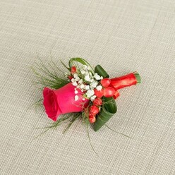 Resting against a pale, sand-toned linen backdrop, this elegant boutonniere looks like it has just been set down on a neatly pressed jacket near a Grove Park dressing room mirror. At its centre, a vivid fuchsia rose commands attention, the petals rich and velvety, spiralling inwards in layers that speak of careful growing and timely cutting. Around the rose, tiny clusters of white baby's breath soften the design, their miniature blooms creating a gentle halo that tempers the intensity of the pink. Among these white flowers sit glistening red hypericum berries, their skin glossy and rounded, lending a hint of warmth and festivity that would suit a winter ceremony or a joyful summer gathering in a nearby Grove Park garden. Slender, feathery fronds of greenery stretch outward, adding airiness and a sense of natural movement, while fuller, darker green leaves are curled and shaped to cradle the rose, giving the small arrangement a sculptural, almost architectural quality. The stems are expertly gathered and wrapped with a smooth, crimson satin ribbon, tied neatly so that no rough ends show, ready to rest comfortably against a suit lapel. The soft, even lighting creates delicate shadows along the textured fabric beneath, highlighting the contrasts between matte petals, shiny berries, and the silk-like sheen of the ribbon. The overall impression is one of poised romance and thoughtful detail, perfect for a groom, best man, or prom guest from the Grove Park area who wants a touch of classic floral elegance that echoes local garden roses and the quiet charm of tree-lined residential streets.