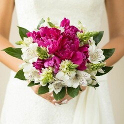 In this intimate photograph, the camera lingers on a bride or wedding guest holding a voluptuous hand-tied bouquet, pressed gently against the subtle folds of a white gown. The arrangement is dominated by full, ruffled peonies in saturated shades of fuchsia and magenta, each blossom layered with petal upon petal until it forms a rounded, luxurious cushion of colour. Nestled between these show-stopping blooms are clusters of white alstroemeria, their slender petals etched with delicate maroon flecks and stripes that draw you in closer, like fine calligraphy on silk. Bright, spiky green buds and textured filler flowers punctuate the composition, their fresh tones and pointed shapes cutting through the softness of the peonies and giving the bouquet a lively, contemporary rhythm. Encircling everything, deep green, glossy leaves arc outward, grounding the piece in nature and adding depth around the edges, much like the mature trees that line the quiet streets of Grove Park. The bride's hands, with neatly manicured, softly pink nails, clasp the stems in a relaxed, almost tender grip, suggesting a moment of calm before the celebrations begin. The background melts into a creamy blur, hinting at lace or tulle but never distracting from the intricate dance of colours and forms in the flowers themselves. The overall feeling is one of confident elegance and heartfelt joy, the kind of bouquet a local Grove Park Florist might create for a couple marrying in a nearby church or registry office, hoping their flowers will stay in the memory long after the last guest has gone home.