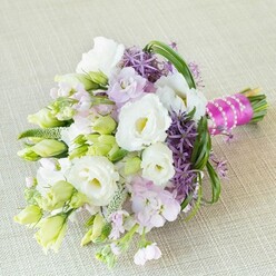 Resting gracefully on a neutral, richly woven beige linen, this hand-tied bouquet by a Grove Park florist exudes refined beauty and fresh optimism, evocative of the gentle breezes near Strand-on-the-Green. Central white lisianthus, softened by ruffled edges and golden-hued centers, form the arrangement's heart, surrounded by fresh white and lime-green buds that hint at coming abundance. Clusters of lavender-pink stock provide an airy, powder-soft layer-their pastel hues creating a harmonious transition to the bold, spiky purple allium heads, which infuse the bouquet with vibrancy reminiscent of the lush borders seen in local riverside gardens. Pale green buds and threaded green fillers interlace through the arrangement, building texture and depth. Long arching strands of bright green bear grass add movement, drawing the eye upward and outward. The bouquet is finished with a striking magenta ribbon, intricately wrapped and set off with a cascade of white pearl strands, reflecting the subtle glamour of Grove Park celebrations. The interplay of creamy white, pastel lavender, and vivid purple makes this a perfect choice for weddings or bridal events in Grove Park-a bouquet that celebrates both unity and the natural beauty of West London's floral traditions.