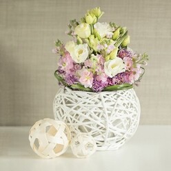 Gracefully set against a subtly textured greige wall evocative of Grove Park's stately homes, this exquisite floral centerpiece radiates both modern elegance and the warmth of the local community. A full bouquet spills over with creamy ivory and pale yellow lisianthus, their gently ruffled petals unfurling like springtime along quiet Grove Park avenues. Punctuating this softness are upright stalks of blush-pink matthiola, adding dynamic vertical lines and a sense of lively abundance, while a foundation of vivid purple filler blooms-likely cultivated from local gardens-grounds the arrangement, offset by a whispering ring of glossy green foliage. The bouquet nestles within a striking, round white vase constructed of intricately entwined painted rattan, its open lattice design echoing the careful craftsmanship that Grove Park Florist is known for. Flanking the vase, two decorative orbs fashioned from pale natural wood mirror the artistry and handmade traditions of the area, their open weave doubling the visual texture and reinforcing the theme of organic beauty. Resting all atop a spotless white surface that reflects gentle, diffused daylight, the entire display breathes with calm, possibility, and an understated sophistication-perfect for a local wedding breakfast or an elegant dinner on nearby Baring Road. Every element here-from the tangle of petals to the play of light-reflects both skilled expertise and a unique Grove Park spirit, inviting memories to blossom among the blooms.