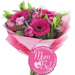 This image captures a joyful new-baby bouquet that looks as though it has just been prepared by a local florist in Grove Park, set against a clean white background where every colour and texture stands out clearly. The focal point is a cluster of radiant fuchsia gerbera daisies, their striking dark centres surrounded by neat whorls of petals that create a strong visual burst of pink. Nestling between these upbeat blooms are delicate pink roses in differing stages of bloom: some are still closed, their petals wrapped protectively around the bud, while others are slightly open, revealing layered interiors that evoke tenderness and affection. Fine wisps of purple filler flowers are scattered throughout the bouquet, providing a soft veil of lavender shade that contrasts gently with the pink and adds dimension. Around and underneath the blossoms, mixed green foliage forms a lush base, with leaves of various widths and lengths giving movement and a fresh, garden-picked feel. The stems are bound together and dressed in tiers of pink tissue paper in complementary tones, artfully arranged into a ruffled, voluminous collar that cradles the flowers. Over this, a shiny layer of clear cellophane encloses the wrap, reflecting small highlights and underlining the bouquet's ready-to-gift finish. Positioned in front of the stems sits a round foil balloon with a bright pink background, playfully decorated with white polka dots. Across its centre, in elegant white script, reads the affectionate message "Mum to Be!" anchored by a small heart design. The overall feeling is one of heartfelt congratulations and gentle excitement, ideal for marking a pregnancy announcement or baby shower gathering in Grove Park, perhaps in a cosy home near the river or a family celebration close to local parks.