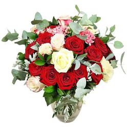 An opulent arrangement of roses stands in a clear glass vase against a perfectly white backdrop, the design radiating romance and careful craftsmanship from a florist who knows how to create impact for homes around Grove Park. The bouquet is generously packed with roses in deep red and bright scarlet, their velvety petals spiralling tightly in some buds and spreading wide in others, like layered silk unfolding in slow motion. Interspersed among these intense reds are creamy white and ivory roses, as well as a few blooms touched with soft, pale pink at the petal tips, lending the composition a gentle glow that tempers the heat of the crimson tones. Delicate clusters of small bright pink filler flowers thread in and out of the roses, offering light, playful notes of colour and extra textural interest around the edges and between the larger blossoms. Encircling the bouquet and reaching slightly beyond the roses, abundant eucalyptus sprigs with rounded, blue-green leaves bring a modern, stylish edge, their subtle silvery cast suggesting the crisp, clean scent often noticed when bouquets are arranged for same-day delivery in Grove Park and the nearby Hounslow borders. Dark green foliage supports the structure, and a few silvery-green leaves break up the deeper tones, helping the design feel balanced, full, and naturally abundant from top to bottom. The vase itself, clear and slightly bulbous at the base before narrowing at the neck, reveals the neatly trimmed stems and fresh water inside, grounding the luxurious arrangement in simple, transparent elegance. With nothing but the white background to compete for attention, the eye is drawn exclusively to the interplay of rich reds, soft neutrals, rosy pink touches, and cool greenery, a visual expression of love, celebration, and thoughtful gifting.
