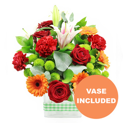 This photograph presents a joyful, garden-inspired bouquet displayed in a modern, clear rectangular vase, the base wrapped with a neat band of light green and white gingham ribbon that evokes a relaxed, kitchen-window feel familiar in Grove Park homes. Above the glass, the flowers form a dense, rounded arrangement where warm reds and oranges are balanced by crisp whites and lively greens. Deep red roses, their petals velvety and tightly packed, serve as romantic focal points. Nestled alongside, red-orange carnations with soft, ruffled heads lend a plush, comforting texture. Bright orange gerbera daisies punctuate the design, each with a dark central eye and a halo of flat petals that draw the gaze outward. Emerging gracefully through the middle, white Oriental lilies add vertical interest; some are fully open, revealing pale pink veins and prominent stamens, while others are still in elongated buds, promising continued flowering. Interwoven throughout are clusters of lime-green button chrysanthemums, their tiny spherical blooms offering a playful contrast in both colour and form. The arrangement is richly backed by glossy deep green leaves and airy sprigs of eucalyptus in cool silver-green tones, which introduce gentle curves and movement. The stems can be seen clearly through the vase, immersed in water that catches the light and hints at freshness. In the bottom right corner, a bright circular orange graphic slightly overlaps the vase and carries the message "VASE INCLUDED" in bold white lettering, making it clear that this is a ready-to-display gift. The overall impression is of an energetic, uplifting bouquet that would brighten a dining table near Chiswick Station, a studio overlooking the Thames, or arrive as a same-day delivery from Grove Park Florist to mark joyful news or heartfelt appreciation.
