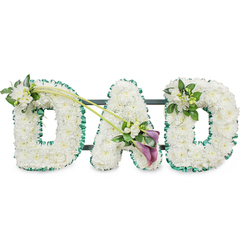 A three-dimensional floral display spelling out "DAD" is the sole focus of this photograph, the letters standing tall on a subtle dark frame that disappears into a white studio backdrop. Each generous block letter is completely covered with immaculate white chrysanthemum heads, meticulously placed so that every petal touches its neighbour, creating a soft yet structured blanket of blooms. The overall effect is one of gentle calm and purity, a fitting choice for a final tribute. Around each letter runs a rich, emerald green satin ribbon, folded into small ruffles that follow the contours and lend crisp definition, echoing the lush greenery and riverside walks that characterise Grove Park. The first "D" carries a small corsage of white freesias and fresh foliage at its upper left, their slender buds and open trumpets adding lightness and a touch of movement. From this point, two delicate green stems sweep diagonally across the arrangement, leading the eye toward the central "A", where an elegant focal spray has been arranged. Here, two deep purple calla lilies, with velvety, trumpet-shaped blooms, are nestled amongst white freesia flowers and vivid green leaves, bringing a subtle but striking contrast to the predominantly white design. The last "D" is finished with a smaller, harmonious cluster of freesias and foliage on its top right curve, mirroring the embellishment on the first letter without overpowering the simplicity of the tribute. The design conveys dignity, tenderness, and gratitude, the sort of carefully composed arrangement that might be delivered by a local Grove Park florist for a funeral or memorial gathering to honour a devoted dad.