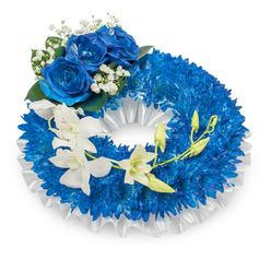 An elegant sympathy wreath in shades of deep blue and crisp white, captured on a clean white background so the arrangement's textures speak clearly. The dominant band is composed of countless small, royal-blue chrysanthemum heads packed tightly together to create a full, tactile ring that feels almost velvety to the touch. Lining the inner circumference and tucked beneath the blooms is a ruffled white satin ribbon with a soft pleat, giving the design a polished, ceremonial edge. At the upper-left curve a cluster of three striking blue roses forms a noble focal point-their petals densely layered in a rich sapphire tone, complemented by tiny sprays of white baby's breath and touches of dark green leaf that add depth and a refined, natural accent. From the mid-left the arrangement carries a gentle waterfall of pristine white orchids-likely Phalaenopsis or Dendrobium-whose delicate, elongated petals arc toward the lower-right, catching daylight with a porcelain-like translucence. Scattered through this cascade are fresh green orchid buds at varying stages of opening, introducing a whisper of new life within a solemn composition. As a local floral tribute, this wreath reflects the calm restraint we often create for services at Grove Park's community chapels and nearby cemeteries, crafted with attention to scent, texture and proportion. It conveys peaceful remembrance and purity-an understated, dignified choice for expressing sympathy and honouring a loved one.
