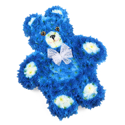 Set on a clean, white backdrop, this floral tribute takes the beloved image of a teddy bear and recreates it entirely in flowers, combining technical skill with deep tenderness. The bear's body is saturated in royal blue, sculpted from hundreds of tightly packed blossoms that resemble small chrysanthemums or hydrangea florets, giving the surface a richly textured, fur-like quality. Light slides softly across the petals, accentuating the curve of the head, the subtle rise of the belly, and the rounded paws, so that the figure feels both dimensional and inviting. Gentle accents of cream and soft butter yellow petals form the rounded muzzle, the inner cups of the ears, and four neat paw pads, these lighter tones lending an air of innocence and warmth. Two polished, dark brown eyes with black centres bring a tender gaze to the face, while a delicate black triangular nose and a slight curved line beneath it create a quiet, comforting smile. A sheer white bow of fine mesh or organza encircles the neck, its gossamer loops and tails lying lightly on the blue flowers like a final affectionate touch. Although the teddy is designed to lie flat, its slight tilt within the frame gives it a sense of gentle movement, as if turned lovingly by hand. In Grove Park, this style of blue teddy bear arrangement is often chosen for a baby boy's funeral, a child's memorial, or as a special tribute delivered to local chapels and homes near Baring Road or Chinbrook Meadows. Crafted with the care of a neighbourhood florist, it offers a symbolic cuddle made entirely from fresh petals, quiet colour, and heartfelt remembrance.