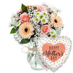 This photograph captures a warm and joyful Mother's Day gift arrangement styled with understated elegance. The scene is set against a spotless white background, which makes every colour appear clear and bright, as if the bouquet were resting on a sunlit table in a Grove Park home. A clear glass vase stands in the centre, its straightforward, cylindrical form giving a clean, modern base for the flowers above. A slim wrapping of rustic twine encircles the neck, forming a small, informal bow that adds a hint of handmade charm. Emerging from the vase, the bouquet forms a soft, rounded dome, filled with pastel tones and crisp whites. Fully opened peach-pink roses reveal tight spirals of velvety petals, and their gentle blush tones are echoed in the peach and light orange gerbera daisies positioned around them. These gerberas have broad, layered petals framing distinctive dark brown centres, providing a focal point within the arrangement. Between the larger blooms, airy clusters of white baby's breath give a subtle mist-like effect, while compact white chrysanthemums with apple-green centres bring structure and a fresh, contemporary note. Deep green foliage is visible throughout, framing the pastel flowers and evoking the leafy character of Grove Park's gardens and riverside paths. Slightly obscured behind the blooms, a small white card peeks through, its partially visible green and pink lettering implying a handwritten "Happy Mother's Day" message. To the right of the vase, leaning forward, a generous heart-shaped foil balloon glows softly. Its bright white background is scattered with tiny metallic gold and silver dots, and at its heart lies a peach-coloured shape bearing the phrase "HAPPY Mother's DAY" in bold black text, with an elegant flourish to the word "Mother's." The composition radiates affection and appreciation, as if ready to be carried down a Grove Park street to surprise a much-loved mum.