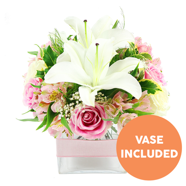 Against a spotless white background, a compact yet luxurious bouquet is shown in full detail, as though freshly arranged at Grove Park Florist for delivery along Burlington Lane. Two prominent white Oriental lilies form the central focus, their broad petals unfurling like gentle waves and revealing dark reddish-brown stamens that add striking vertical lines. Around them, clusters of pink roses are gathered closely, some in firm, tightly wrapped buds and others in open, ruffled blooms that suggest tenderness and affection. Among the roses weave slender stems of alstroemeria, in soft tones of pink and white, their delicate, elongated petals tipped and streaked with hints of yellow and fine speckles, bringing a sense of intricate artistry. Tiny white baby's breath is scattered throughout, creating a light, airy veil that softens the transitions between the larger flowers and lends the bouquet a dreamy, cloud-like quality. The entire arrangement is framed by a rich selection of green foliage: broad, shiny leaves reflect the light, while finer, feather-like fronds introduce texture and a sense of natural movement, reminiscent of the varied greenery along the Thames towpath near Grove Park. All the stems are carefully arranged inside a modern, square, clear glass vase, where the clean lines and visible stems contribute to the overall feeling of freshness and order. A pale pink satin ribbon wraps horizontally around the vase's middle, adding a graceful finishing touch that echoes the soft rose tones above. In the bottom right corner of the image, a circular orange label with bold white capital lettering reads VASE INCLUDED, clearly highlighting that the container is provided with the flowers. The image conveys a mood of calm sophistication and quiet joy, ideal as a centrepiece for an intimate dinner or a thoughtful gift delivered locally.