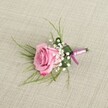 A single blush pink rose boutonniere rests on a softly textured light beige linen surface, photographed from a gently elevated, eye-level angle so you can almost feel yourself standing above a dressing table in a Grove Park townhouse just before a wedding. The rose is semi-open, its velvety petals spiralling out from a tight centre of pale, pastel pink into deeper, more romantic pink tones along the softly curled outer edges, suggesting a bloom cut at perfect freshness. Clusters of airy white baby's breath surround the base of the rose, each tiny flower creating a cloud-like halo that lightens the design and adds that classic, timeless touch so many local couples around Grove Park choose for their church and registry office ceremonies. Fine, wispy blades of bright green, grass-like foliage fan out delicately behind the rose, giving the boutonniere an ethereal frame and adding movement, as if a light breeze from nearby Grove Park itself had brushed through the greenery. A broader, darker green leaf is neatly folded around the lower portion of the stems, forming a structured cradle that keeps everything compact and elegant. At the base, a narrow pink satin ribbon, echoing the soft tones of the rose, is wrapped smoothly around the stems and tied into a small, tidy bow with very short tails. The gentle, even lighting casts subtle shadows, emphasising the layered petals, the crisp edges of the leaf, and the woven linen texture below. This detailed image captures a classic pink wedding boutonniere ready to be pinned to a groom's or prom guest's lapel, an understated yet romantic finishing touch for celebrations across Grove Park and neighbouring streets.