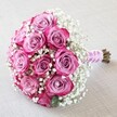 Balanced atop a gently textured, light cream linen, this bouquet evokes the classic charm and understated romance Grove Park is known for. Around a dozen roses form the heart-lush, with petals unfurling from a subtle lavender-pink at their centre to bright magenta and fuchsia on the outermost tips, each blossom pristine and artfully shaped. Tucked between and around these vivid roses, snow-white gypsophila is arranged to provide a dreamy softness, their tiny blooms floating like delicate clouds above the main flowers. The bouquet is tied with care, a smooth pink ribbon holding the stems together and winding seamlessly into a spiral of small, glowing white pearls-details that call to mind the craftsmanship of local Grove Park florists. Just below, trimmed stems in earthy greens and browns add to the feeling of just-picked freshness. The whole scene is lit with gentle, diffused daylight, emphasizing each velvety petal and the ethereal structure of the baby's breath. With its rounded, full-bodied profile and harmonious palette, this bouquet feels both modern and timeless-the perfect floral touch for weddings in the Grove Park area or any occasion celebrating love, unity, and enduring elegance.