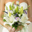 A masterful wedding bouquet that feels at once refined and relaxed, composed to glow in soft daylight and to travel easily from bridal prep to ceremony. Large, star-shaped white lilies form the heart of the design, their velvety petals opening in layers so the eye moves from one generous bloom to the next. Interspersed are lisianthus in pale white with delicate purple edging; their ruffled shapes introduce understated drama and a cool violet note that amplifies the lilies' purity. Small veronica spikes and clusters of baby's breath add slender vertical accents and a misty texture, while fine grass-like foliage and broader green leaves provide a living frame that breathes around each flower. The arranger has staggered buds and open flowers to suggest movement-some lilies still promising future bloom, some lisianthus curling softly-so the bouquet reads as both composed and alive. In the subdued sunlight typical of a Grove Park morning, creamy whites take on a tender warmth and the violet edges hold a jewel-like clarity; scent drifts softly, a floral sweetness tempered by fresh green undertones. This bouquet was carefully balanced for weight and hand-feel, arranged so it sits comfortably in the bride's grip without losing visual impact. As a Lewisham florist familiar with small, heartfelt ceremonies near Grove Park station and the Nature Reserve, Grove Park Florist designed this piece to suit intimate church weddings and local registry-room vows alike, and its calming palette makes it adaptable for both classic and contemporary bridal styling.