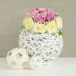 This photograph captures a low, tightly formed arrangement of roses in harmonious pastel tones - blush pink paired with creamy vanilla-yellow - each bloom arranged to create a plush, rounded silhouette that reads both romantic and restrained. Interspersed among the roses, feathery clusters of baby's breath (Gypsophila) provide a soft, cloud-like texture that frames the bouquet and gives a sense of lightness, as if the blooms might float from the vase. The container is a sculptural orb of white-washed rattan, its woven strips crossing in an open lattice that offers glimpses into the inner stems and lets subtle daylight from a nearby window play across the petals. Two coordinating decorative orbs made from pale, natural wood sit to one side on the same bright white surface, one nearer the vase and the other slightly forward, forming an intentional but relaxed vignette. The neutral beige backdrop carries a faint horizontal linen texture, lending a domestic warmth that suits both contemporary and traditional interiors. The scent here would be quietly floral and fresh, the tactile contrast between the roses' velvety petals and the rattan's smooth, cool weave reinforcing the arrangement's handcrafted quality. Imagined for a wedding table, an anniversary, or a tasteful living-room focal piece, this arrangement reflects thoughtful floral design and seasonal care - the kind of composition I might prepare for a morning delivery across Grove Park, leaving a soft, fragrant impression on a recipient returning from the high street or Grove Park Station.