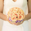 There is a gentle poetry to this bridal bouquet-a hymn of apricot warmth and twilight-lilac accents arranged with careful symmetry so the roses read like gathered sunbeams. Each apricot rose unfurls with a creamy centre and delicate outer petals that curl in soft spirals; they are set in a compact cluster, framed by a neat ring of purple-tipped lisianthus whose cool tones lift the composition and add a refined contrast. Between these primary blooms, a fine lattice of tiny white filler flowers gives the whole arrangement an airy, almost floating quality, the sort of detail that sings quietly beside bridal lace.
Light falls across the bouquet in a soft, flattering way, drawing attention to petal texture and the subtle veins that mark each rose-an effect that photographs beautifully in both late-morning sunshine and the warm glow of reception rooms. The scent is described as fresh and lightly sweet, a classic rose perfume tempered by green notes from the foliage, lending a calm, uplifting atmosphere to the bride as she walks down the aisle. The bouquet's compact scale and balanced silhouette make it ideal to be paired with vintage lace or a modern, minimalist gown.
Prepared with local care by Grove Park Florist and suitable for same-day delivery across Grove Park, Lewisham and nearby Catford, this arrangement is crafted to be the quiet centrepiece of a wedding day: intimate, composed and full of promise. It's both a keepsake of the moment and an object that complements vows, photographs and the little rituals that make a ceremony feel truly personal.