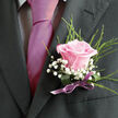 This image shows a finely crafted wedding boutonniere pinned neatly to a dark, tailored lapel, the kind of detail you might notice on a groom stepping out of a car near Grove Park's tree-lined streets. At its centre is a single pink rose, petals layered in a gentle spiral, their blush tone soft and even, like a quiet sunrise. The rose is fully formed but not open to excess, balancing elegance and freshness. Around it, fluffy clusters of white gypsophila sit closely together, each tiny bloom creating a starry texture that softens the edges of the design. Wisps of feathery greenery emerge from behind, slender and upright, giving a sense of lightness and drawing the eye upward along the lapel. The boutonniere's stems are wrapped in a lilac satin ribbon that hugs them tightly, the smooth material finished in a tidy fold near the pin. The ribbon's cool lavender shade lies between the pink of the rose and the depth of the suit fabric, gently echoing tones often chosen in wedding colour palettes. Subtle natural light touches the petals, making their texture appear almost touchable, while the dark suit cloth offers a crisp contrast that lets every element stand out clearly. This piece feels personal and intimate, as though prepared carefully for a ceremony just off the main avenues of Grove Park, where family and friends will gather. It's a small symbol of love and commitment rendered in flowers and fabric, designed to be worn all day yet remembered long afterwards.
