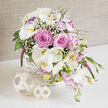 On a smooth white surface, a luxurious basket of wedding flowers glows softly, as though ready to be carried into a quiet ceremony in Grove Park. The basket itself is woven from white wicker in a tight, cross-hatch pattern, offering a clean, textured base that contrasts beautifully with the lush, spilling blooms. Center stage, large blush-pink roses are fully open, their velvety petals layered in generous spirals that give depth and a sense of tender abundance. Sweeping around them, white Phalaenopsis orchids extend gracefully beyond the rim, their broad, delicate petals and yellow-flecked centers casting subtle shadows that hint at their sculptural form. Intermixed among these key flowers, white lisianthus blossoms unfurl in soft tiers, some with just a kiss of lavender at their edges, echoing the cooler undertones of the arrangement. Airy sprays of baby's breath are threaded through the bouquet like a fine veil, adding volume without weight and imparting a dreamy, cloudlike effect. Clusters of smaller, soft pink flowers tumble with the longer stems, enhancing the asymmetrical, cascading silhouette. Rising in contrast, slender spikes of deep magenta or purple Veronica draw the eye upward and add an elegant vertical rhythm. Green foliage in varied shapes and tones nestles between the blooms, grounding the pastel palette with fresh, natural depth, like foliage gathered from a sheltered Grove Park garden. In front of the basket, two decorative, pale rattan or wood-shaving spheres rest on the tabletop, one larger, one smaller, their open lattice echoing the woven basket. The softly textured beige backdrop and gentle lighting give the whole scene a warm, refined calm, making this design ideal as a centerpiece for a Grove Park wedding reception or an unforgettable romantic delivery to a local home.