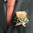 A study in restraint and warmth, this boutonniere features a flawless peach rose mounted against a charcoal lapel, its petals layered like soft porcelain and bathing the composition in coral-pink light that mirrors the adjacent tie. Circlets of white gypsophila encircle the central bloom, creating a cloud-like texture that contrasts with the rose's velvety surface; the tiny star-shaped flowers scatter highlights and lend a whimsical innocence. Wisps of fresh green foliage - a slender curled leaf and delicate fern-like fronds - provide structural rhythm and a verdant freshness reminiscent of early-morning dew. The base is finished with a pale peach satin ribbon tied into a modest bow, the ribbon's sheen repeating the tie's tone and holding the stems with quiet precision. When photographed against a charcoal suit, the colours read clearer, the peach glowing more vividly and the minute details of the gypsophila and foliage coming forward. There's a subtle scent, a soft rose perfume mingled with cut green stems, that suggests care taken in the arranging. Locally, this style of boutonniere is a favourite for Grove Park weddings and celebratory services from Baring Road to the high street; crafted fresh in the morning and prepared with steady hands, it reflects the attentive approach of neighbourhood florists. At Grove Park Florist we see this combination frequently - an elegant, sentimental accent made to be pinned close to the heart on an important day.