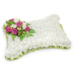 A rectangular pillow-shaped memorial crafted entirely from a dense carpet of small, pristine white chrysanthemums that creates a soft, cushion-like surface and reads as quiet comfort. The tightly packed blooms give the piece a pillowy texture that looks almost textile under soft studio light, the petals catching a gentle sheen while a faint shadow beneath the form defines its three-dimensional presence. On the upper left corner sits a concentrated spray of colour: several deep magenta-fuchsia roses with petals unfurling toward the viewer, their velvety faces offset by tiny clusters of pure white freesias or similar buds that lend a delicate fragrance note in the imagination. Sprays of glossy green foliage and slender fern fronds cradle the roses, adding depth, contrast, and a natural framework that draws the eye across the arrangement. A neat pleated trim of light green satin ribbon runs around the pillow's edge, slightly visible beneath the white chrysanthemum mass and giving a subtle flash of fresh colour and formality. The overall palette - immaculate white, lush green, and rich magenta - reads as peaceful, respectful, and tender. Presented against a clean white background, the tribute maintains a calm, uncluttered dignity appropriate for funeral or graveside services, offering a visual language of rest and remembrance. As a local Grove Park floral design, it feels suited to quiet services in nearby chapels or green spaces such as Chinbrook Meadows, where families seek a serene, understated symbol of lasting love.