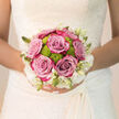 This bridal bouquet, held close against the intricate lace of a wedding dress, captures the emotional stillness just before a ceremony in Grove Park, when everything is ready and time seems to pause. At the centre, generously petalled roses in shades of lavender-pink and muted mauve sit proudly, their velvety surfaces catching the light with a gentle sheen. Each rose feels full and reassuring, like a promise. Tucked between them are lively clusters of bright green chrysanthemums, their tidy, button-shaped blooms providing a zing of freshness and a contemporary note, echoing the lush green spaces that define Grove Park. Around the edges, creamy-white blossoms and slender buds form a soft, cloud-like halo, toning down the contrast and bringing the colours into a harmonious blend. The bouquet is hand-tied in a neat, rounded shape that fits perfectly in the bride's hands, the stems gathered in a way that offers both comfort and poise. Light falls softly over the arrangement, emphasising the gentle interplay of colours-dusty pinks, cool greens, and warm whites-while leaving the background understated so the flowers remain the focus. You can almost breathe in the scent: classic rose with hints of green stems and fresh foliage, the kind of fragrance that stays with you long after the day is over. Crafted with the understanding of a local Grove Park wedding florist used to styling flowers for riverside venues and intimate church aisles, each element of the bouquet has been chosen to last through photos, vows, receptions, and late-night toasts. It feels both grounded and romantic, a floral reflection of joy, hope, and the start of a shared life.