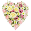 A tender heart-shaped floral arrangement is shown in full view against a bright white background, its colours soft yet expressive, its form clearly defined. The heart is packed tightly with flowers, so that the entire surface appears plush and complete, without empty spaces. At the core, several large cream-to-buttery yellow roses open in graceful spirals, their smooth, layered petals suggesting warmth, care, and the gentler memories held close by those left behind. Encircling them, clusters of pale pastel pink carnations with frilled, velvety petals build the main body of the heart, creating a gentle outline that feels like a visual embrace. Among the carnations, white spider chrysanthemums rise up, their slender, radiating petals surrounding bright green centres, offering delicate texture and moments of crisp contrast. Pockets of deep magenta-pink lisianthus are placed thoughtfully throughout the design, their bell-shaped blooms and softly ruffled edges introducing a richer hue that brings emotional depth and quiet dignity. Small leaves and hints of green foliage peek between the blossoms, enhancing the sense of freshness without disturbing the overall softness of the heart's shape. At the very tip, a narrow pink ribbon glimmers subtly, hinting that this piece has been lovingly prepared as a funeral, sympathy, or memorial tribute, perhaps destined for a service in Grove Park, Chiswick, or nearby Brentford. The mood of the arrangement is calm and consoling, as if it has been created to sit silently beside a family in their grief, offering beauty, comfort, and a final gesture of affection when words feel too fragile.