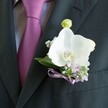 A close-up photograph focuses tightly on the left lapel of a groom's charcoal suit, showing a white orchid buttonhole resting against a finely tailored jacket and a rich mauve tie, as if captured just outside a Grove Park registry office. The fabric of the suit is a deep charcoal grey, almost black, with a soft, matte weave that absorbs light and makes the flowers stand out with painterly clarity. Beneath the lapel edge, a smooth tie in deep magenta-plum glows against a pale grey dress shirt with a discreet textured pattern, the knot sitting crisp and confident at the collar. Pinned neatly at the lapel, a large Phalaenopsis orchid is the undisputed focal point, its broad, satiny petals a pure, luminous white that seems to hold a cool sheen, like porcelain warmed by gentle afternoon light. At its centre, a small flare of yellow and soft orange in the throat adds warmth and life. Around this single, sculptural bloom, tiny clouds of lilac blossoms and white gypsophila tuck in close, their fine, airy petals giving a sense of movement and softness, reminiscent of spring borders along the paths in Grove Park. Slender sprigs of deep green foliage frame the orchid and echo the natural curve of the lapel, giving the buttonhole structure and depth. A small bud of pale green orchid stands just above the main bloom, hinting at new beginnings. Everything is gathered and finished at the base with a narrow lavender satin ribbon, tied into a tidy bow that mirrors the tie's tone, as though coordinated by a florist who understands both fashion and flowers. The mood is quiet, romantic elegance: a moment of poised anticipation before vows are spoken, captured in a single, meticulously arranged orchid boutonniere.