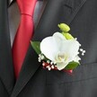 A close-up photograph focuses on a single wedding boutonniere pinned neatly to the lapel of a dark charcoal grey suit jacket, the fine woven texture of the fabric clearly visible beneath the flowers. At the heart of the design sits a large white Phalaenopsis orchid, its broad, velvety petals fanning out in a soft curve and framing a delicate yellow-and-white lip at the centre. Tucked just above the orchid are two small, round lime-green blooms, shaped like tiny chrysanthemums, their compact, cushiony texture adding a playful contrast. Around the orchid, sprigs of cloudlike white baby's breath scatter tiny blossoms, creating a light, airy halo against the deep cloth. A handful of polished, rich green leaves peek out from behind, giving the boutonniere a fresh, garden-picked feeling. The stems are bound in a slim, deep red satin ribbon that echoes the strong red of the solid satin tie beside it, which lies over a pale grey and white micro-checked dress shirt. The red tie cuts a striking line through the composition, hinting at a groom or groomsman dressed for a refined Grove Park wedding, perhaps heading from a nearby church to a reception by the common. Soft, even lighting glides over every surface, highlighting the orchid's gentle sheen, the slight gloss of the ribbon, and the clean edges of the suit's pocket just beneath the flowers. The overall mood is one of understated sophistication and quiet romance, the kind of carefully considered detail a local florist might pin in place just before the ceremony, ensuring the wearer steps out feeling composed, elegant, and ready to celebrate.