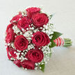 In this photograph, a timeless bouquet of red roses and white gypsophila is presented with calm elegance, evoking a wedding morning in Grove Park. The roses dominate the centre of the design, their petals a deep, velvety red, spiralling from tight hearts into open, rounded blossoms that almost seem to glow in the soft light. Each bloom sits close to its neighbour, forming a smooth, dome-like silhouette that looks both full and carefully ordered. Threaded delicately throughout are clusters of baby's breath, their fine, branching stems studded with countless small, white flowers, like a gentle veil laid over the stronger reds. The contrast of textures is striking: the substantial, almost plush quality of the roses set against the feathery, cloud-like gypsophila. Subtle flashes of green foliage sit around the base and peep between stems, drawing the eye outward and grounding the design in nature, much like the leafy surroundings of Grove Park and nearby Chiswick. The stems themselves are neatly aligned and bound into a tidy handle, swathed in ribbon and finished with a trim of pearl-like beads that lend a touch of refinement, as if the bouquet has been crafted at Grove Park Florist for a couple saying their vows near the river. The natural lighting is soft and flattering, giving the reds a rich, romantic depth and the whites a luminous, almost ethereal glow. You can almost imagine holding the bouquet, feeling the cool stems against your palm and catching a whisper of sweet rose fragrance as you move. Everything about the image suggests commitment, tenderness, and a moment suspended in time, ideal for weddings, vow renewals, or intimate celebrations across W4.