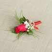 A single fuchsia rosebud boutonnière is laid diagonally across a soft, light beige fabric that looks like finely woven linen, the neutral background allowing every petal and leaf to stand out clearly. The rosebud itself is tightly furled, a deep, saturated pink that leans toward magenta, with velvety petals spiralling in on themselves to suggest a bloom just about to open. Nestled close to its base are tiny tufts of white baby's breath, each miniature blossom forming a cloud-like halo around the rose and bringing a gentle, airy contrast to the strong colour of the central flower. Behind and around the rose, slender sprigs of bright green, feathery foliage reach outward in soft arcs, giving the boutonnière a light, almost whimsical silhouette, as if it has just been gathered from a garden near Grove Park and arranged with a careful, practiced hand. Deeper green leaves sit nearer the stem, anchoring the piece with a sense of structure and depth. All of the stems are neatly gathered and wrapped in a glossy red satin ribbon, wound firmly yet delicately, with the tail forming a tidy, decorative loop that hints at the care of a professional florist. The lighting is even and bright, picking up the gentle sheen of the ribbon and the subtle texture of the fabric while highlighting the freshness of the petals and foliage. The overall impression is one of polished romance and understated elegance, perfect for a groom or prom guest walking through Grove Park on a clear, celebratory day, boutonnière pinned to a lapel and ready for photographs and special memories.