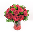 A lavish, tightly clustered bouquet of deep crimson red roses fills the frame, each bloom perfectly formed and velvety, as if freshly gathered from a morning delivery into Grove Park. The roses are packed closely together, their petals spiralling from tight, secretive centres into broad, open cups that catch the light and reveal layers of plush texture. The red is intense and saturated, somewhere between ruby and wine, evoking romance, passion, and an almost film-like sense of drama. Around and between the roses, rich dark green foliage-likely Ruscus or a similar glossy greenery-creates a vivid contrast, its smooth, slightly waxy leaves framing each flower head and giving the bouquet a full, luxurious shape. All of this is anchored in a translucent ruby-red glass vase with a spiralled or twisted design that subtly ripples across its surface. Light glances off the etched pattern, creating delicate highlights, while the rose stems, visible through the coloured glass, add depth and structure. The vase flares gently at the top, with a solid, grounded base that makes the arrangement feel stable and composed, as though ready to take pride of place on a Grove Park dining table or windowsill overlooking the local green. Set against a spotless white background, the rich reds and greens seem to glow, every flower at peak freshness. The composition suggests an unmistakably romantic gift, ideal for anniversaries, proposals, or heartfelt declarations, the kind of bouquet whose soft fragrance might linger across an evening, turning an ordinary London night into a cherished memory.
