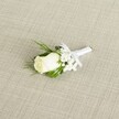A single white rose boutonniere is laid diagonally across a softly textured, light beige fabric, the weave resembling natural linen you might find in a Grove Park townhouse near the station. At the heart of the image, a creamy ivory rosebud sits tightly furled, its velvety petals curving inward with a gentle promise of full bloom. The base of the bloom shows a hint of pale green sepals, grounding the whiteness with a touch of natural freshness. Around the rose, clusters of tiny white baby's breath form a delicate halo, their cloud-like blossoms adding a whispery, romantic softness that recalls spring ceremonies in nearby Chiswick or along the leafy streets of Grove Park. Slender, deep green leaves and a few more feathery, fern-like fronds cradle the flowers, creating a rich contrast that emphasizes the purity of the rose. The stems are neatly bound in smooth white satin ribbon, the wrap perfectly even and finished with a modest, symmetrical bow just below the flower heads, clearly designed to sit neatly on a lapel for a wedding or formal event. The lighting in the photograph is gentle and diffused, with no harsh shadows, just a calm glow that highlights the satin sheen of the ribbon, the subtle curves of each petal, and the airy texture of the gypsophila. This boutonniere feels intimate and timeless, the sort of carefully considered finishing touch a groom or groomsman might choose for a classic Grove Park wedding, capturing the quiet excitement and polished elegance of the day in one small, beautifully crafted piece.
