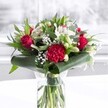 A luxurious bouquet in a clear cylindrical glass vase stands proudly at the centre of the image, as if just placed on a bright kitchen table in a Grove Park townhouse. The water in the vase is crystal clear, revealing a carefully spiralled bundle of neatly trimmed green stems, each one aligned to show the florists' precision and care. Above the waterline, the arrangement bursts into a rich palette of deep red and pure white, framed by abundant greenery. Velvety crimson roses open in gentle spirals, their outer petals slightly unfurling to suggest romance and devotion, while a large ruffled red carnation sits near the front, its crimped edges and densely layered petals adding drama and texture. Threaded between the passionate reds are creamy white alstroemeria with slender, slightly flared petals etched with soft speckles, along with chubby white lily buds held tightly closed, hinting that the bouquet will keep changing and opening over the coming days. Clouds of tiny white gypsophila, like scattered stars, fill in the gaps, giving the whole design a soft, airy halo. Around the base, broad, glossy dark green tropical leaves are wrapped and gently curled, forming an elegant collar that grounds the design and reflects the light. Finer eucalyptus-style foliage and pointed green leaves stretch upwards and outwards, creating depth, movement, and a natural garden feel. The bouquet is photographed on a pristine white surface, with a softly blurred, light-filled background that could be a window overlooking Grove Park's leafy streets. The gentle, even lighting catches every petal and leaf, creating a feeling of freshness and quiet celebration, suitable for an anniversary dinner, a Mother's Day surprise, or simply to brighten a local home or office with heartfelt affection.