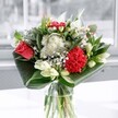 A contemporary mixed bouquet sits in a clear glass vase on a sunlit surface, captured as if in a quiet home near Grove Park station. At the heart of the arrangement, a single coral-red rose and a velvety crimson carnation draw the eye, their tightly layered petals rich with texture and depth. Around them, clusters of crisp white daisy-like chrysanthemums and tiny button blooms create a soft, clouded halo, their centres a gentle yellow that adds warmth to the overall palette. Creamy white lilies, some open and some still in smooth, tapered buds, rise slightly above the other flowers, lending a sculptural feel and a sense of anticipation, as though the bouquet will look a little different with each passing day. Deep emerald foliage, glossy and carefully shaped, curves around the base in broad arcs, cradling the flowers and giving the design a polished, modern silhouette. Light from a nearby window, reminiscent of a calm morning along Marvels Lane, falls across the glass and leaves, highlighting every vein and petal edge, and casting delicate shadows that suggest movement. The whole composition feels fresh and uplifting, like a cool breath of air after rain in Grove Park, perfect for celebrating a birthday, sending warm wishes, or simply brightening a living room with an air of thoughtful elegance and quiet joy.