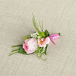 This boutonniere showcases a beautifully formed pink rose, its petals layered like delicate silk, moving from a richer blush at the centre to the faintest, almost translucent pink at the edges. Nestled close to it are light alstroemeria blossoms, their pale pink petals and softly golden throats decorated with fine streaks of deeper pink, introducing a subtle sense of pattern and rhythm. Tiny white gypsophila blooms cluster between the larger flowers, shaping a fine, airy halo that brightens the overall silhouette. Around the flowers, slim green grasses and tidy leaves provide a fresh, structured frame, echoing the gentle greenery found along Grove Park's quiet lanes and nearby riverside paths. The stems are drawn together and carefully wrapped in a glossy pink ribbon, smooth and slightly reflective, adding a neat, polished finish that hints at the formality of weddings and milestone celebrations. Resting against softly textured fabric, the piece appears ready to be lifted and pinned to a lapel, its curves and lines designed to sit comfortably against a tailored jacket. In the mind's touch, you could sense the velvet surface of the rose, the cool firmness of the stems beneath the ribbon, and the crisp tip of each leaf. This boutonniere feels well suited to a groom, usher, or guest setting out from Grove Park towards a ceremony in nearby Chiswick or along the Thames, offering a quiet yet confident statement of tenderness, care, and the importance of the day.
