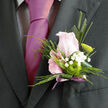 Captured in close detail, this boutonnière feels like a quiet love letter in flowers, ready to be pinned on in a Grove Park home just before the first guests arrive. At the centre lies a delicate, blush-pink rose, the bloom held at that perfect moment of opening when each petal gently lifts away from the next, full of softness and subtle texture. Encircling it are light sprays of white gypsophila, dozens of miniature florets combining to create a soft, cloud-like frame that softens the edges and brings an almost dreamlike quality to the design. Beside the rose, a rounded green button chrysanthemum provides a strong counterpoint, its crisp, compact petals and vivid colour adding both structure and a fresh, contemporary twist. Slim leaves and neat greenery trace graceful lines up the boutonnière, some arcing gently outward while others hug the central stems, echoing the natural calm of the tree-lined roads around Grove Park station. The stems are finished with a length of mauve ribbon, wrapped smoothly and securely, with a small trailing end that catches the light in soft lavender tones. The overall impression is of thoughtful simplicity: romantic but not overstated, traditional yet gently modern, perfect for a wedding by the river or a formal celebration in nearby Chiswick or Kew. You can almost imagine the wearer glancing down as the pin is fixed, feeling the cool satin, the faint fragrance of rose, and the reassuring presence of something beautiful crafted especially for them. It is a piece that speaks of care, of time taken, and of the quiet artistry that turns fresh flowers into a moment remembered.