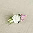 An elegant wedding boutonnière lies on a diagonal across a softly textured, oatmeal-beige fabric, the weave reminiscent of fine linen you might find in a Grove Park townhouse. At its heart sits a fully opened white lisianthus bloom, its many ruffled petals curling outwards in loose, cloud-like layers, the centre softly shaded with a hint of cream. Just below and to the left, two slender lisianthus buds in pale green and ivory rest close to the main flower, capturing that quiet moment before they unfurl. Threaded between them are tiny sprays of white gypsophila, each blossom no larger than a pinhead, creating a misty halo around the focal flower and adding a sense of lightness and air. Fine, grass-like blades of deep green foliage arc gracefully from behind the bloom towards the lower left corner of the image, giving a gentle sense of movement, while a single broader, darker leaf sits snugly behind the lisianthus, grounding the design. The exposed stems are neatly gathered and wrapped low on the right with a vivid fuchsia satin ribbon, tied into a small, neat bow whose silky sheen catches the light and introduces a joyful pop of colour against the neutral fabric and white petals. The lighting is soft and even, like a calm afternoon in a Grove Park garden, revealing every petal edge and subtle texture without harsh shadow. The overall mood is one of simple, modern romance, a small yet carefully considered floral detail ready to be pinned to a groom's lapel for a local ceremony, perhaps after a quiet walk through nearby Grove Park with the photographer capturing those first married moments.
