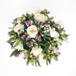 From an overhead perspective, this bouquet appears as a tranquil circle of soft colour and texture, resting on a spotless white background that highlights every detail. At the centre, creamy white roses cluster tightly, their velvety petals unfurling in elegant spirals that move from cool white edges into subtle ivory and delicate peach within, like the gentle glow of late afternoon light over Grove Park's quiet streets. Some roses are fully opened, revealing their intricate hearts, while others are still partly closed, suggesting grace and continuity. Woven through this central mass of blooms are slender alstroemeria stems, mostly in bud, their tapered forms washed with deep purple and lavender at the tips, progressing into muted greens and slightly pinkish bases. These buds, caught just before opening, add a feeling of expectation and ongoing life. Wisps of white baby's breath are scattered throughout, tiny round blossoms creating a fine, cloud-like layer that softens the stronger shapes and adds a sense of stillness. Deep green foliage surrounds and supports the arrangement, broad leaves with a gentle gloss forming a protective cradle around the more delicate flowers. The bouquet holds a compact, spherical shape, speaking of careful, deliberate design by a Grove Park florist mindful of creating something both beautiful and comforting. The light falling on the bouquet is even and soft, avoiding harsh contrasts and instead offering gentle shadows that accentuate texture. Together, the creamy roses, purple buds, and airy Gypsophila convey sympathy, peace, and quiet reassurance, making this arrangement fitting for sending condolences or placing in a serene corner of a home in Grove Park.