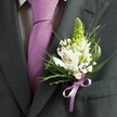 A close-up, high-angle photograph shows the upper torso of a man dressed for a formal occasion in Grove Park, his attire captured in crisp detail. He wears a charcoal grey suit jacket with a sharply tailored lapel and smooth shoulders, the fabric matte yet refined under soft natural light. Beneath the open lapel sits a pale dress shirt with a fine grid pattern, its cool tones perfectly offset by a sleek lavender tie, neatly knotted and falling straight down the centre of his chest. Pinned to the left lapel is the true focus of the image: a carefully crafted wedding buttonhole featuring a tall, pale green spiky bloom, likely an unopened hyacinth or Muscari, rising gracefully to give height and structure. Around it, clean white alstroemeria blossoms open with painterly streaks of deep maroon or plum, their petals slightly curved and their golden stamens just visible at the heart. Tiny clouds of white gypsophila, or baby's breath, drift between the larger flowers, softening the arrangement with an airy, romantic feel reminiscent of a quiet morning walk through Grove Park's tree-lined streets. Slender, grass-like blades weave through broader, darker foliage, adding movement and a fresh, garden-picked charm. At the base, a satin lavender ribbon is tied into a small, precise bow, echoing the colour of the tie and drawing the eye to the skilled craftsmanship. The overall impression is of calm sophistication and understated joy, a groom or groomsman pausing for a moment before stepping out into a day of vows, photographs, and celebration just a short stroll from Grove Park station.