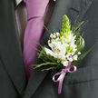 This photograph centres on the chest and lapel of someone dressed for a significant celebration, with the camera lingering on the interplay between fine tailoring and careful floristry. The suit jacket is a deep charcoal grey, its lapel sharply pressed and subtly stitched, giving a sense of craftsmanship you might associate with a groom preparing along a quiet Grove Park side street. Beneath, a pale shirt with an almost invisible dark micro-check pattern peeks out, adding depth without distracting from the smooth, solid lavender tie that falls straight and true. The tie's satin finish gently reflects the light, its soft purple tone immediately echoed by the delicate ribbon that secures the boutonniere. That boutonniere is a refined composition of white alstroemeria with their distinctive inner markings-fine reddish-brown speckles and slender lines that lead the eye to the warm yellow centres-arranged so that each petal feels airily layered but precise. Rising above them, a slender, conical green flower bud introduces a fresh vertical line, suggesting new beginnings, while scattered tufts of white baby's breath lend a light, cloudlike softness. Around the blooms, a mix of narrow, spiky greenery and soft, feathery fronds creates a pleasing contrast against broader, darker green leaves that cradle the design. The lilac satin bow at the base ties everything together, literally and visually, mirroring the tie's tone and giving the impression of a carefully planned colour story, perhaps chosen with a local florist's guidance for a Grove Park ceremony, prom, or anniversary dinner. The overall impression is one of cool elegance and tender anticipation, as if the wearer is poised just moments before walking into a memory they will carry for years.