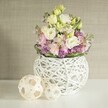 An elegantly composed wedding table arrangement, inspired by Grove Park's relaxed sophistication, sits enveloped in gentle natural light. The centerpiece features a generous bouquet overflowing with multi-layered, ruffled white and creamy yellow lisianthus, each petal unfolding in a soft embrace, reminiscent of blossoms found in neighbourhood gardens near Chiswick Station. Above and among them rise blush-pink stock blooms, lending a subtle fragrance and vertical grace, while the base is animated by clusters of lively purple filler flowers nestled among lush green foliage. All are cradled within a distinctive orb-shaped vase, meticulously crafted from ivory-toned painted rattan in an open-weave design-its artisanal, airy texture adding a touch of modern Grove Park artistry to the classic floral mix. Resting upon a spotless white surface, the arrangement is accompanied by two textile-inspired decorative spheres of natural rattan, echoing both the vessel's craftsmanship and the tactile pleasures of a Grove Park home. The scene's soft, diffused illumination and the creamy textured wall in the background transform the flowers' subtle palette into a vision of calm and new beginnings. This inviting display not only sparks ideas for wedding centrepieces or seasonal home styling in Grove Park, but also suggests the thoughtful touch and care that only a local florist can provide.