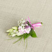 Laid on a subtly woven, neutral fabric that could easily be part of an elegant suit laid out before a big day in Grove Park, this boutonnière captures the charm of understated romance. At the centre, creamy ivory buds are held just before full bloom, their petals soft and slightly curved, giving the impression of something precious and carefully timed. Nestled beside them are pale blush-pink flowers, each petal thin and silky, catching the soft light in a way that gives them a gentle, luminous glow. Tiny tufts of baby's breath cluster around the main blooms, their small white florets adding a cloudlike delicacy that makes the central flowers appear even more refined. Wispy blades of fresh green grass and narrow foliage weave through the composition, adding light movement and a whisper of wild garden energy, reminiscent of a stroll through the tree-lined streets off Grove Park Road on a mild spring morning. Broader, glossy leaves cradle the flowers from behind, shaping the boutonnière and giving it a sense of structure that would sit neatly on a groom's lapel or a smart jacket for a school prom. The stems are bound tightly at the base with a pale rose-pink satin ribbon, its surface smooth and slightly reflective, tied in a tidy knot that speaks of care and craftsmanship. You can almost sense a soft floral fragrance rising from the buds-a subtle blend of fresh, clean sweetness, more evocative than overpowering. The overall mood is one of quiet celebration and personal significance, a small floral detail prepared with the same level of thought someone might give to choosing their venue or favourite local restaurant in Grove Park for their gathering.