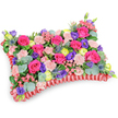 A vibrant pillow-shaped floral tribute photographed from a gently elevated, oblique angle against a pure white background, the composition feeling like a soft, visual embrace. The surface is densely layered with hot pink roses-buds and partially unfurled blooms whose velvety petals catch cool studio light-balanced by clusters of pale shell-pink carnations that introduce a delicate, ruffled texture. Scattered among them are deep purple lisianthus (eustoma), their bell-like forms pushing subtle shadow and depth into the arrangement, while cheerful lemon-yellow ranunculus punctuate the palette with sunny discs of layered petals. Several silver-blue eryngium thistles provide a striking, spiky counterpoint, their bristled heads lending an architectural edge and a cooler tonal contrast to the warmer pinks. Rounded, silvery eucalyptus leaves and assorted rich green foliage weave through the massed flowers, softening edges and lifting the composition with their matt, fragrant surfaces. The entire cushion rests on a distinctive pleated red satin ribbon base, the ruffled border tracing the rectangular pillow silhouette and adding a refined, tactile frame. Scent is implied-powdered rose and eucalyptus-while the overall mood reads as celebration within remembrance: lively color choices that honour memory with warmth rather than solemnity. The craftsmanship shows careful symmetry and dense, supportive mechanics beneath the blooms, the kind of considered construction a local florist in Grove Park might assemble for a funeral or memorial service. Seen from this angle, the tribute reads as both an intimate offering and a public sign of love and comfort, suitable for placing at a chapel, graveside, or wake in and around Grove Park and Lewisham.