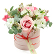 A stylish pink hatbox brimming with flowers is centred against a crisp white background, drawing attention to its soft pastel tones and carefully balanced design. The cylindrical box is a gentle blush-pink with a smooth, matte appearance, wrapped in a narrow coral-red satin ribbon that meets at the front in a neat bow. Suspended from the bow is a small, cream-coloured tag inscribed in green script, suggesting a handwritten note or the name of a trusted local florist, ready to deliver along the leafy streets of Grove Park and toward the riverside paths. Above the rim, the bouquet forms a rounded, luxuriously full dome, with blooms spilling slightly over the edges in a relaxed yet polished way. At the centre lies a striking pale pink rose, fully open and showcasing a deeper pink spiral at its core, which immediately draws the gaze. Surrounding it, tidy white chrysanthemums shaped like daisies, each with a bright green centre, offer fresh, clean contrast, evoking the charm of a cottage garden. Interspersed among them are graceful white alstroemeria, their elongated petals marked with fine speckles, adding a touch of sophistication. Pockets of deeper colour appear in small clusters of vibrant fuchsia or deep pink flowers, likely spray roses or lisianthus, giving lively pops of intensity. The flowers are framed with an assortment of greenery, including rounded, silver-green eucalyptus leaves, darker foliage, and fine stems tipped with tiny buds that enhance the arrangement's texture. The mood is romantic, joyful, and gently celebratory, making this hatbox ideal for birthdays, anniversaries, or quiet gestures of appreciation within Grove Park, where understated elegance and thoughtful details are especially cherished.