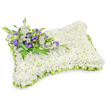 A tender, rectangular sympathy pillow crafted from hundreds of small, pristine white chrysanthemum heads sits like a soft resting place; the dense cluster of blooms forms a plush, cloud-like surface that quietly suggests peace and purity. The pillow's gently rounded corners and slightly raised, puffy profile are visible beneath the floral mass, and soft studio light casts a subtle shadow beneath it that gives the tribute three-dimensional presence. Around the entire perimeter a delicate band of light green satin ribbon is pleated into neat, overlapping loops, the fresh lime tone providing a gentle, uplifting contrast to the sea of white and neatly defining the pillow's edges.
Diagonally across the upper-left corner, a composed spray of accent blooms and foliage balances the design. Crisp white lisianthus with their ruffled petals nestle beside deeper purple lisianthus - some open, some still in bud - their cool violet notes offering a respectful, dignified counterpoint to the white base. Slender fern fronds and glossy, darker green leaves weave through the cluster, and a broader wrap of deep green foliage secures the stems into a tidy, natural bind. There is a faint, clean scent of chrysanthemums and fresh greenery, the kind of quiet fragrance you expect at a memorial service.
This arrangement is made with the calm deliberation of an experienced florist and is suited to graveside tributes, funeral services, or a home memorial. Locally sensitised to families in Grove Park and neighbouring Downham and Lee, the piece reads as a comforting, visual embrace - a floral expression of condolence that speaks where words may fall short.