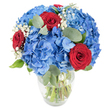 A luminous, hand-arranged bouquet sits in a simple clear glass vase, photographed against a clean white backdrop so every colour and texture reads clearly. The composition is dominated by dense, rounded heads of sky-blue hydrangea whose many tiny petals cluster together like a cool summer cloud; their soft, almost satin surfaces catch the light and give the arrangement a calm, expansive feel. Tucked through that blue cloud are three deeply saturated red roses, their velvety petals still tightly cupped, each one a small, intense point of heat against the cool hydrangea field. Fine sprays of white baby's breath (Gypsophila) drift between the larger blooms, creating whisper-thin highlights that lift the overall silhouette and add an ethereal, delicate rhythm. Muted green eucalyptus leaves, round and slightly silvered, weave through the bouquet, offering a soft herbal scent you can almost imagine and a visual counterpoint that deepens the palette. The transparent vase reveals fresh green stems submerged in water, a reassuring sign of recent craftsmanship and careful conditioning. Bright, natural light sculpts gentle shadows and brings out the layered textures - the hydrangea's dense mass, the rose's plush spirals, the baby's breath's airy points, and the smooth eucalyptus. The arrangement reads as celebratory and composed, equally suited to a birthday table, an intimate anniversary, or a patriotic display where blue and red take on added meaning. Imagine this piece warming a dining table after a walk through nearby Grove Park or as a thoughtful gift for a neighbour in the local high street community.