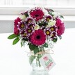 A vibrant mixed bouquet sits proudly in a clear, textured glass vase, its rounded shape framed by a soft, bright white background that suggests a sunlit window in a calm Grove Park home. Deep fuchsia and magenta gerbera daisies, each with a velvety dark centre, form the bold heart of the arrangement, their wide petals radiating out like little suns. Around them, multi-petalled chrysanthemums in rich purple with tips fading to white cluster together, each bloom revealing a cheerful yellow centre that glows in the gentle light. Nestled between these statement flowers are creamy white roses, some fully open with layered, satin-like petals, others still in tight buds, hinting at blooms yet to come and adding a refined, romantic note. Fine sprigs of white baby's breath thread delicately through the design, creating a cloudlike softness and giving the bouquet a light, airy texture, while deep green foliage at the base and edges provides grounding structure and a lush backdrop for the brighter tones. The squat jar-style vase is cinched at the neck with rustic twine tied in a neat bow, and a small white rectangular tag with a simple floral logo hangs from it, suggesting this is a thoughtfully chosen gift rather than a casual bunch. The overall effect is joyful yet elegant, as if prepared by an experienced local florist for same-day delivery around Grove Park, perfect for celebrating a birthday, saying thank you, or brightening a windowsill overlooking the leafy streets near the station.