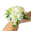 A delicate wrist corsage rests elegantly on a fair-skinned wrist against a clean white background, the image composed to emphasise texture, purity and quiet celebration. At its heart sits a creamy ivory rose, petals unspooling with a velvety softness that catches the light and gives the bloom a gentle, warm glow. Nearby, a larger trumpet-shaped white flower - likely alstroemeria with a slightly ribbed form - opens to reveal a pale green-yellow throat, introducing a subtle, luminous contrast that draws the eye downward through the design. Scattered around these central blooms are compact, verdant green buds, some tightly closed and promising future blossoms, while sprays of tiny white Baby's Breath create a mist-like softness that lifts the arrangement away from the skin. A small cluster of white, daisy-like florets with bright golden centers nestles close to the wrist, offering a sweet, classic punctuation to the composition. Broad, glossy deep-green leaves frame and cradle the flowers, their smooth, waxy surfaces amplifying the fresh tones and providing a rich backdrop to the pale petals. The whole is secured by a refined multi-strand pearl bracelet: uniform, lustrous beads that wrap comfortably and add a tactile coolness against the arm. The lighting is soft and neutral, highlighting petal texture and pearlescent shine while preserving the arrangement's understated elegance. Imagined for weddings, prom nights or formal dances, this corsage suggests quiet joy and tradition - a piece a Grove Park bride or prom-goer might choose when seeking something timeless and softly romantic for a meaningful day.